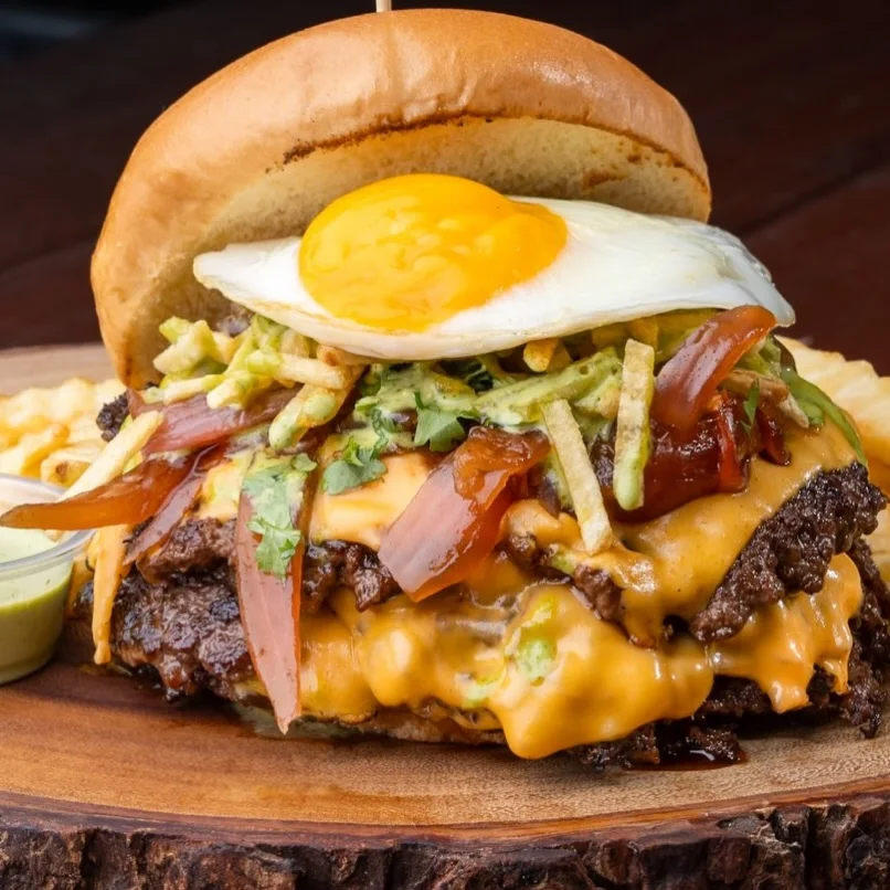 Burger topped with a fried egg, guacamole and potato sticks on a wood board