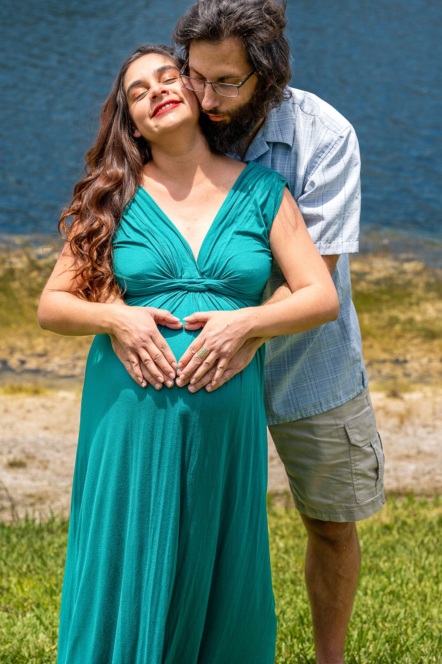 Expecting couple embracing by the water as he kisses her forehead