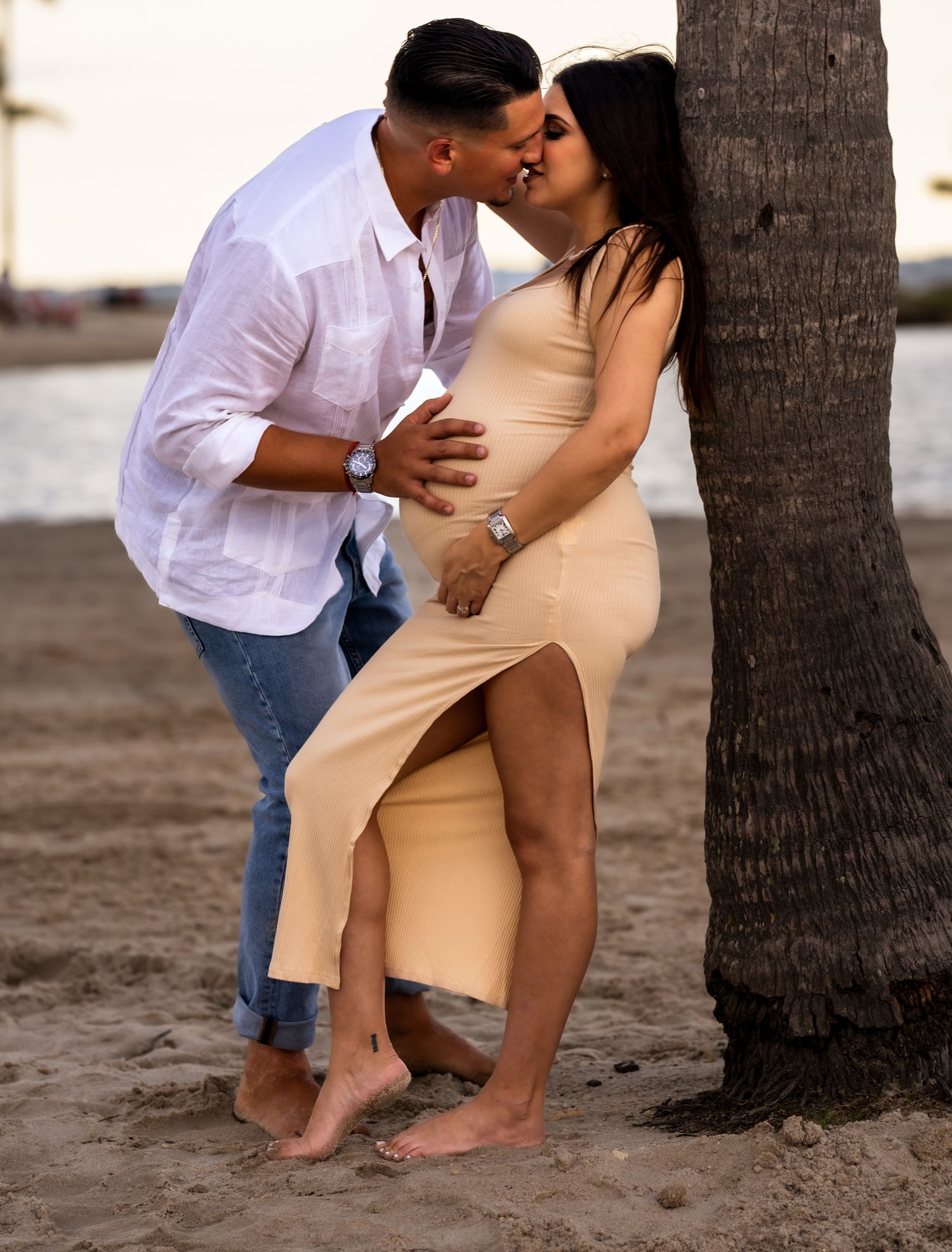 Expecting couple kissing on the beach at dusk, her tan dress catching the evening light