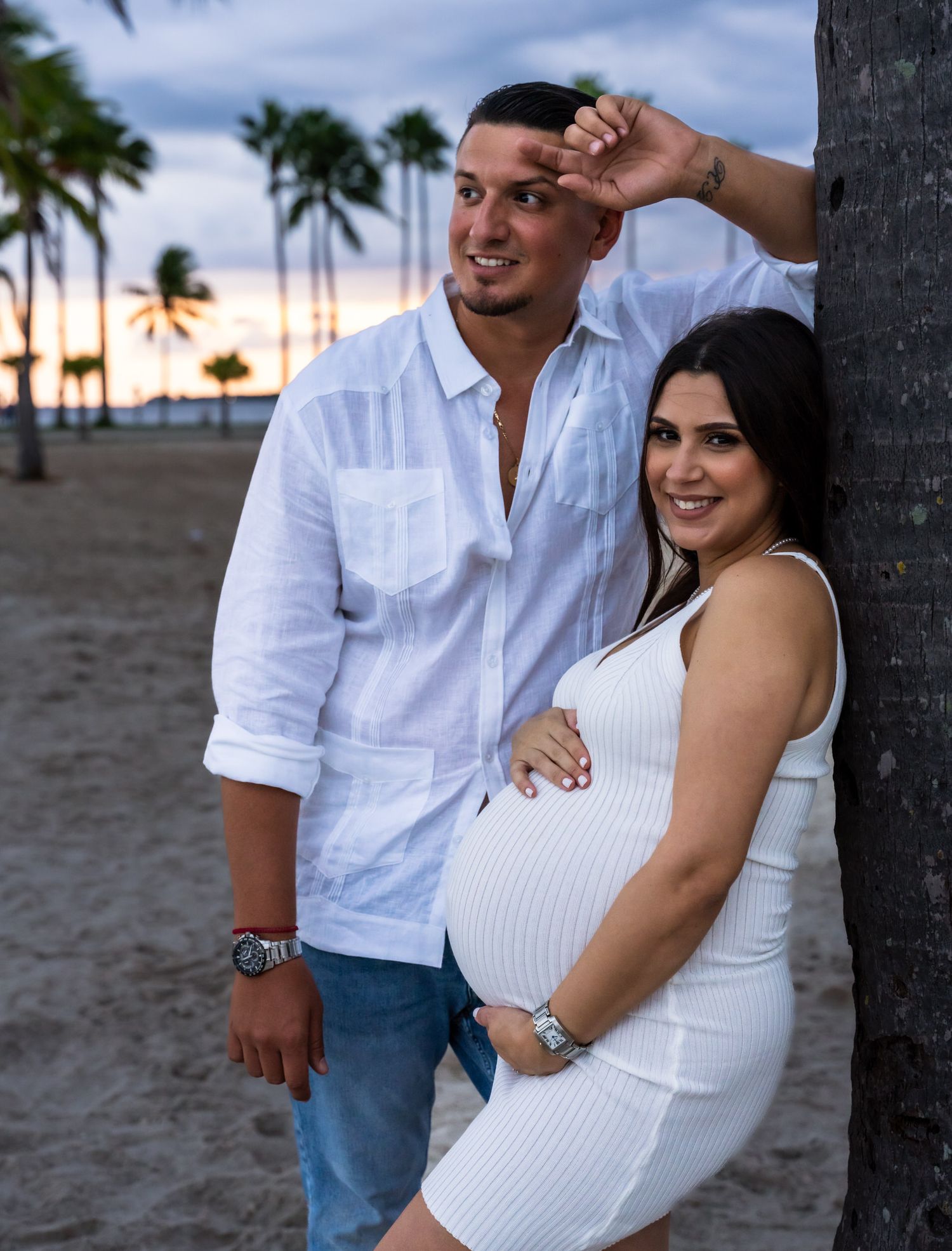 Expecting couple laughing on the beach, he leans against a palm tree beside her