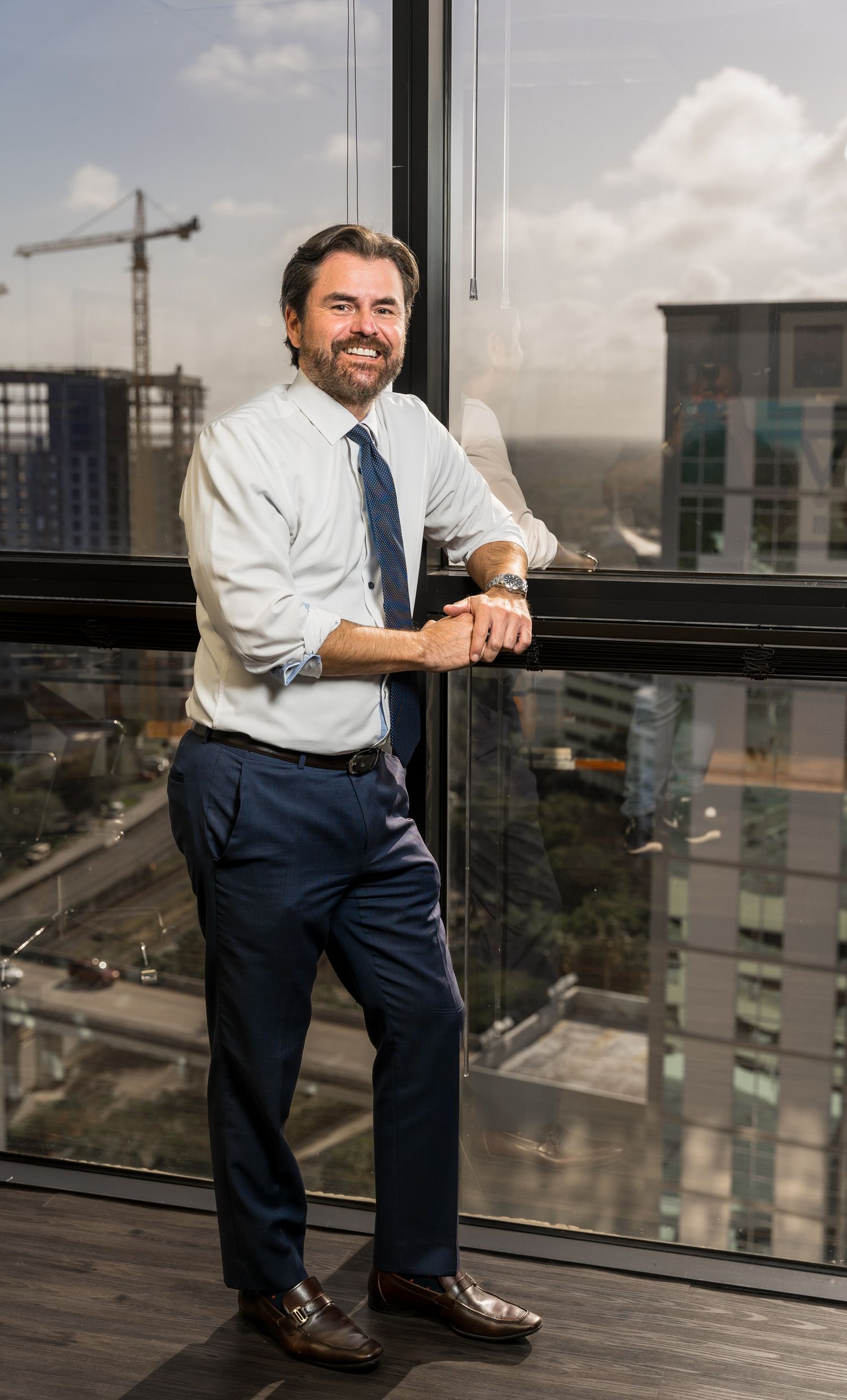Executive portrait leaning against a floor-to-ceiling office window with a city view