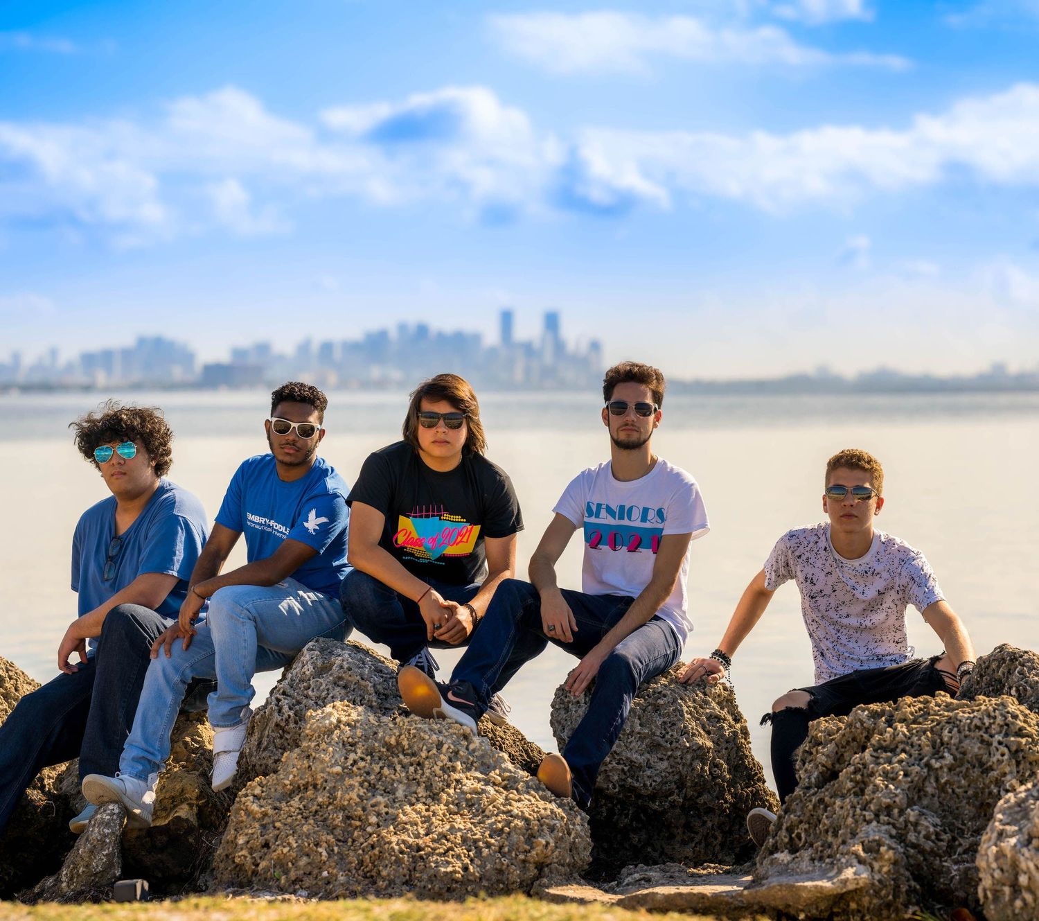 Un grupo de seis amigos sentados juntos en las rocas del malecón de Miami con los edificios detrás