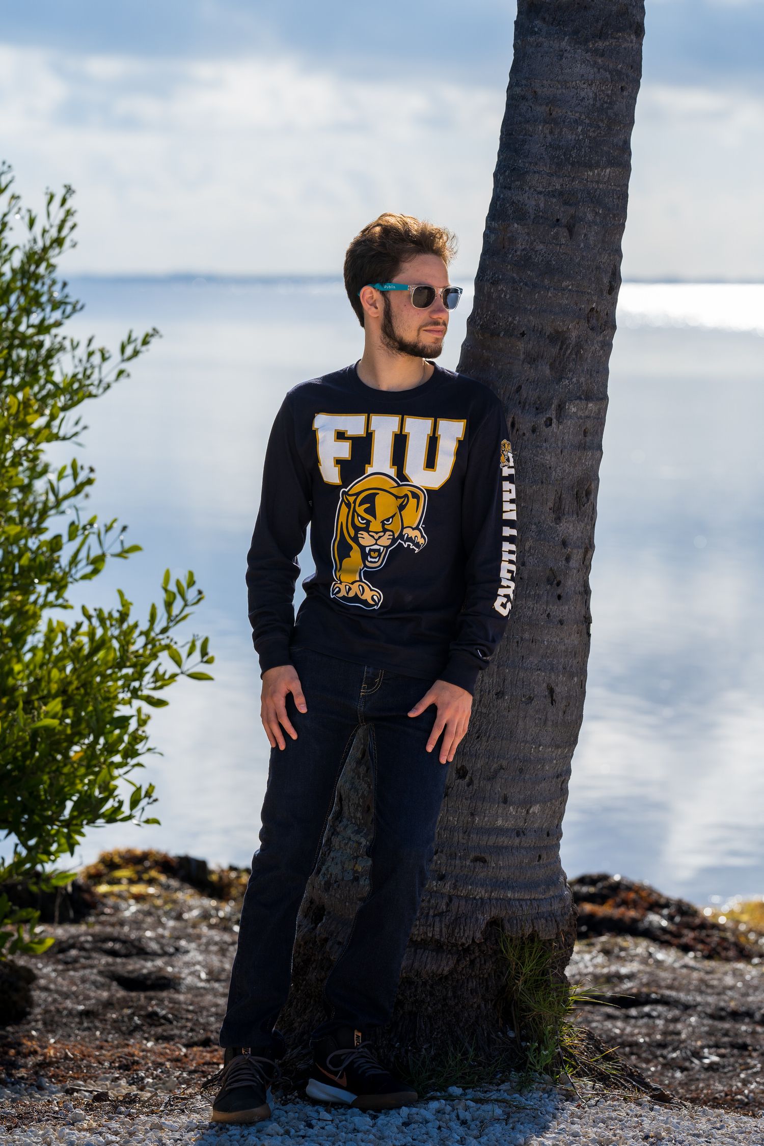 Graduate in an FIU sweatshirt leaning against a palm tree by the water