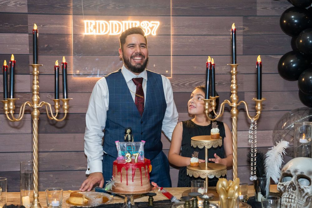 Host couple posing behind an elegant birthday dessert table with gold candelabras and a cake