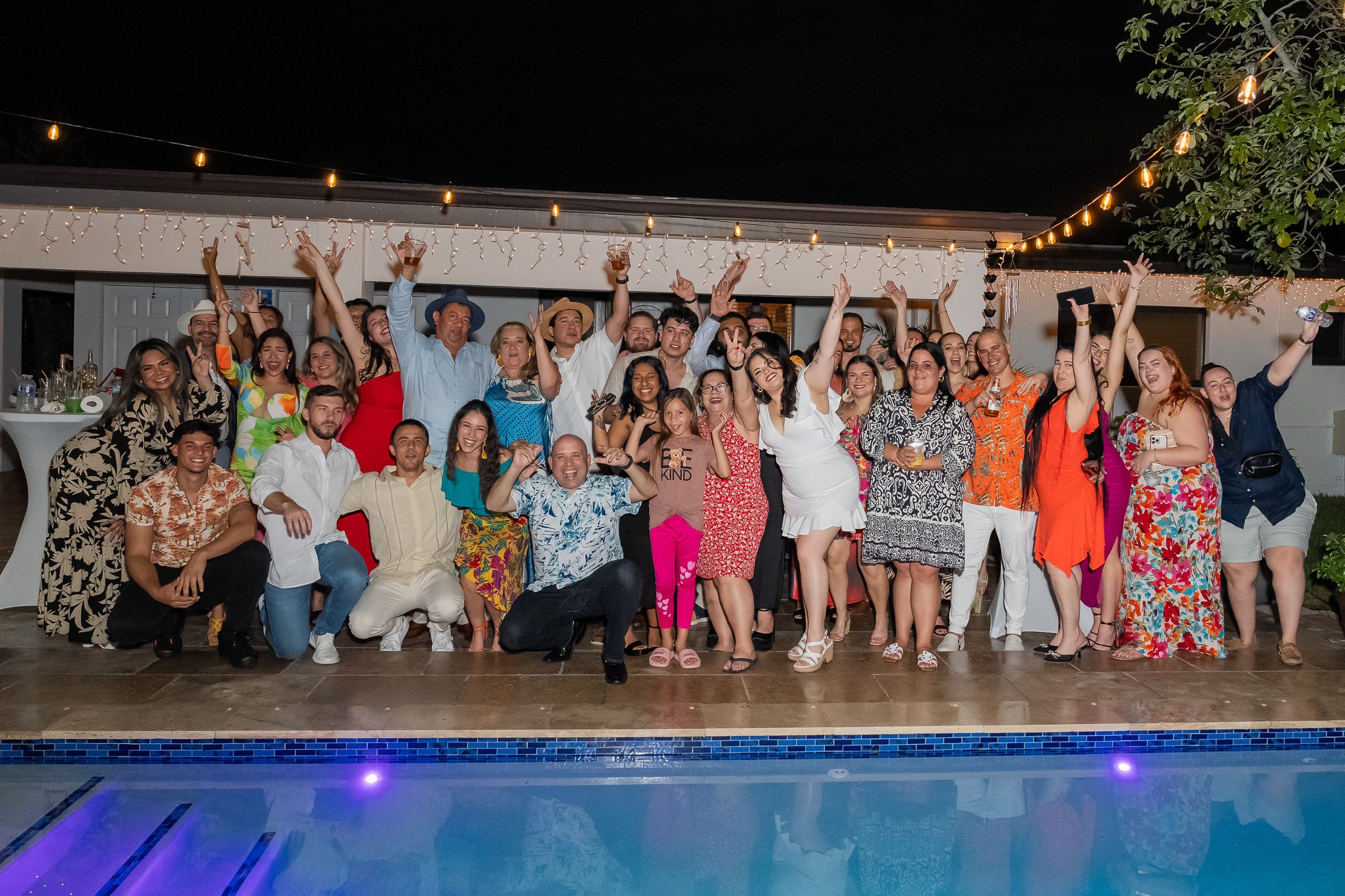 Un grupo grande de invitados celebrando junto a la piscina en una fiesta de noche, con guirnaldas de luces encima
