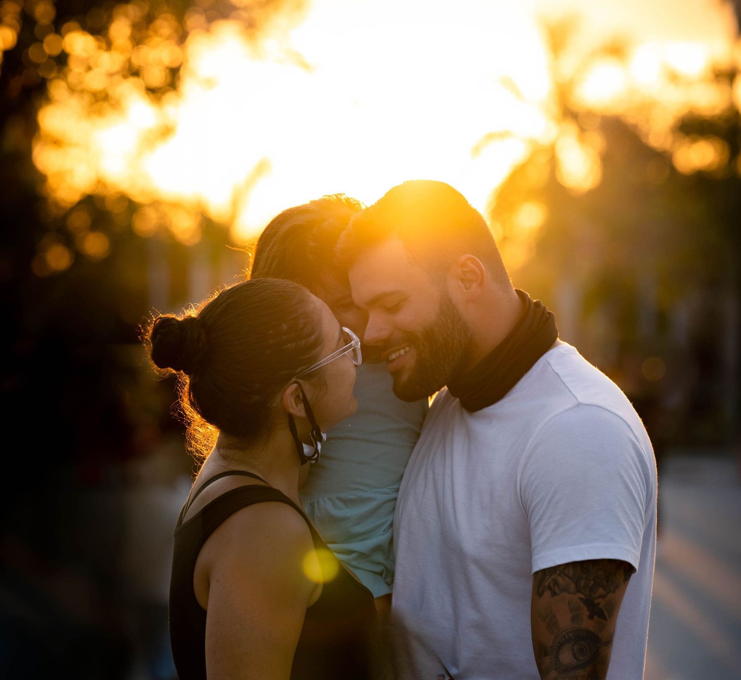 Backlit sunset portrait of parents with their foreheads resting against their toddler's between them