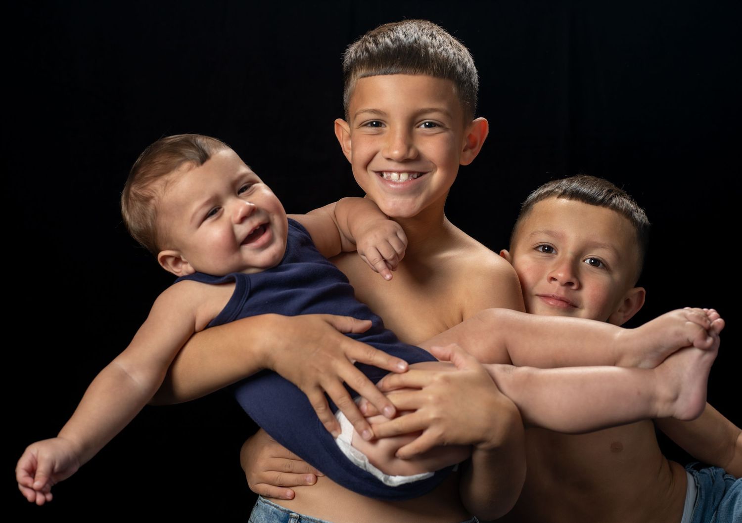 Three brothers in a studio portrait, the oldest carrying his baby brother while his other brother holds on