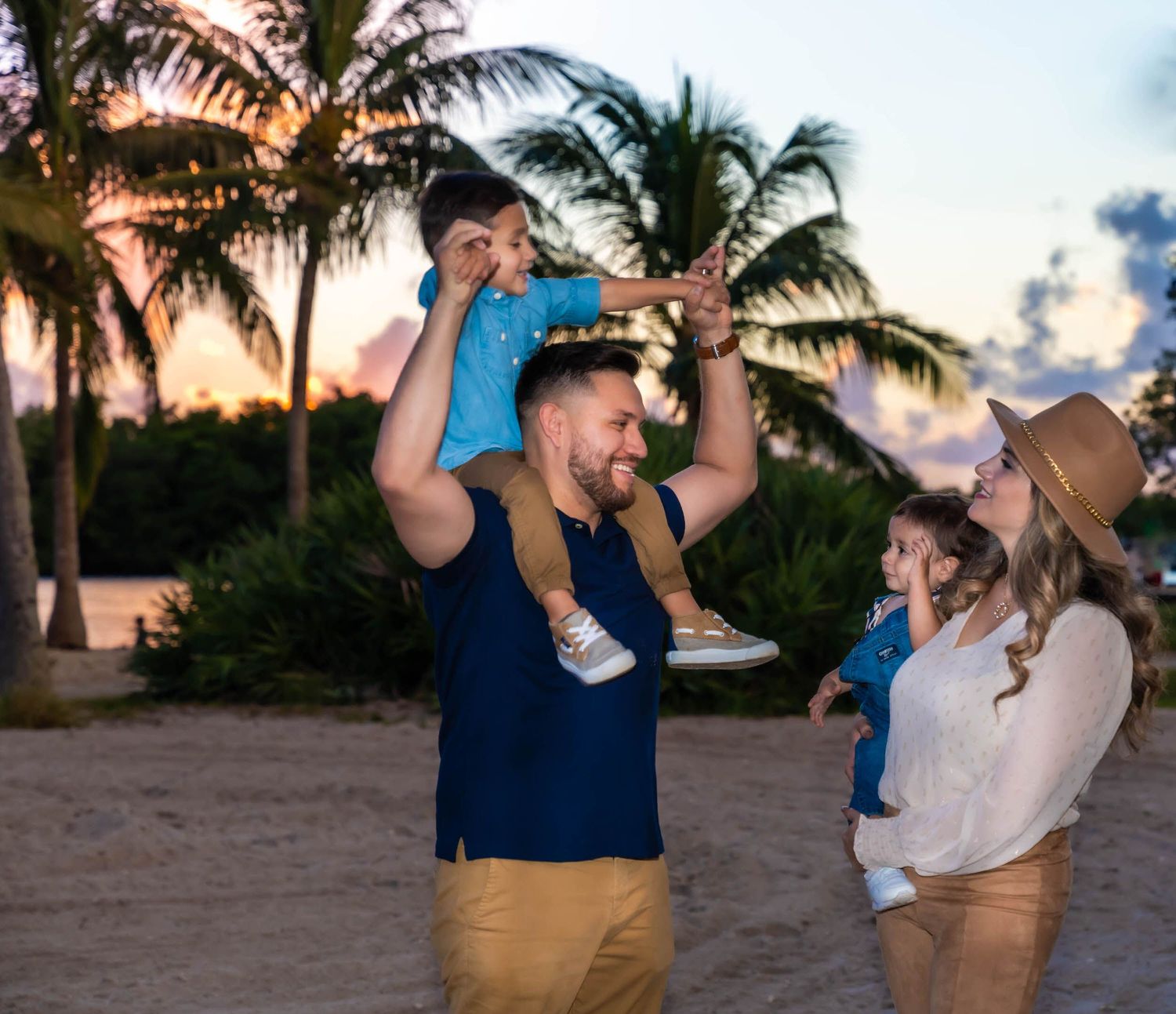 Father swinging his son overhead by the hands while the mother holds their toddler, palm trees at sunset