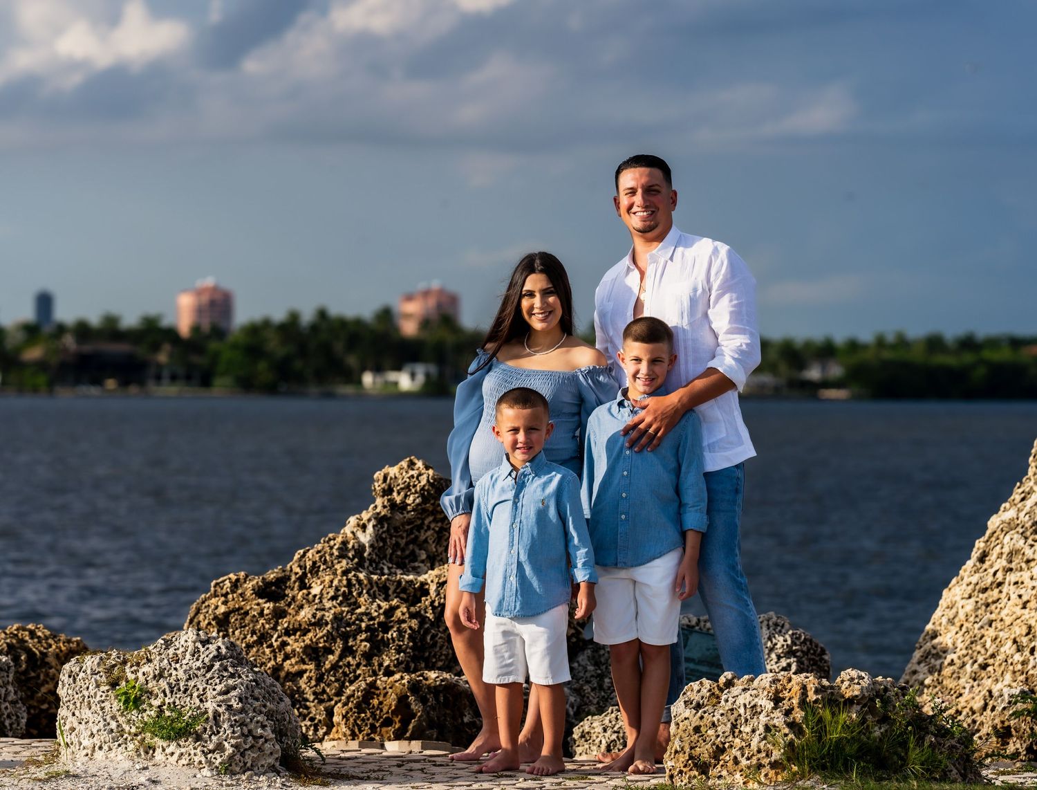 Family of four standing on coral rocks by the water, two young sons in chambray shirts between their parents