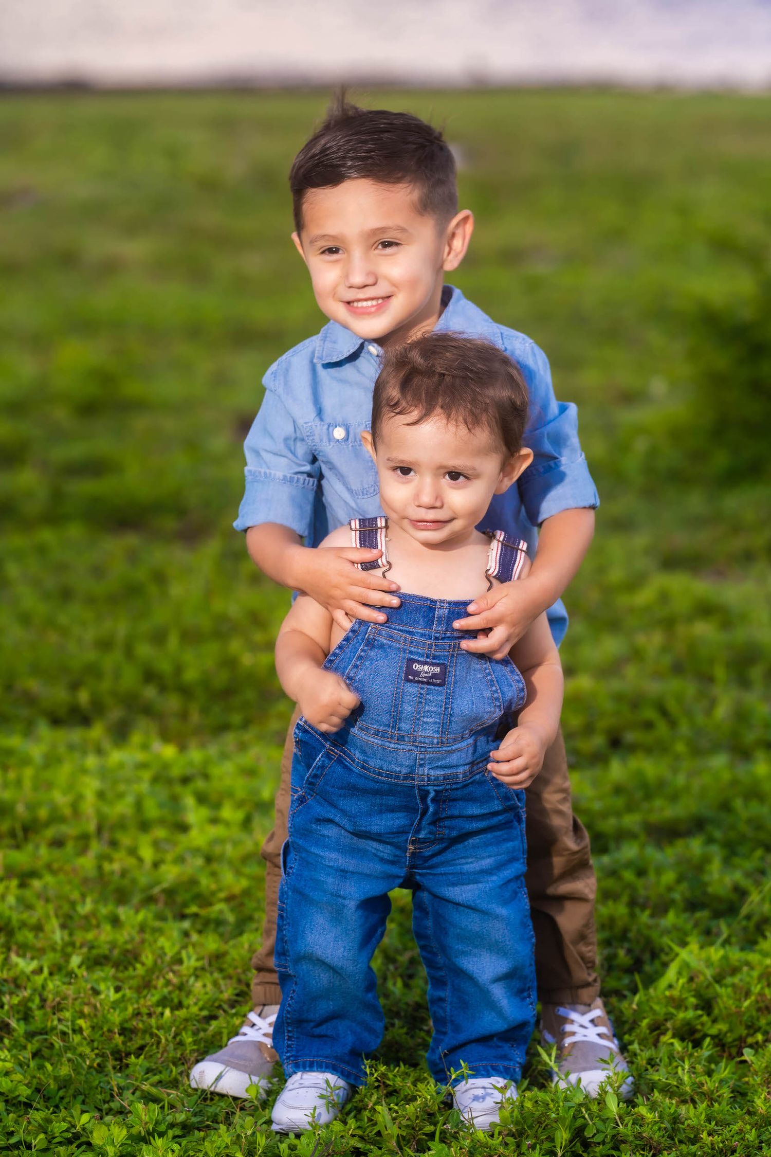 Older brother with his hands on his baby brother's shoulders, standing in a grassy field