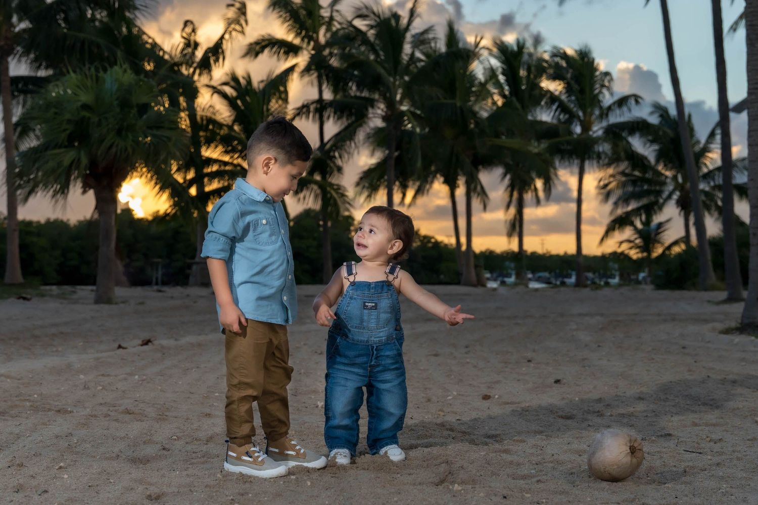 Two young brothers talking on the beach at sunset, the younger one pointing at a coconut on the sand