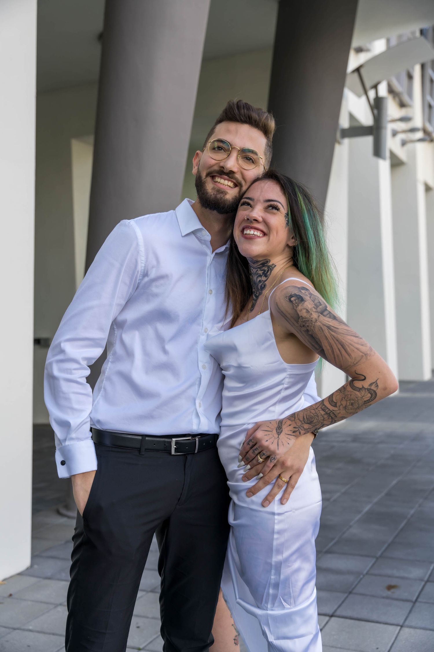 Newlyweds embracing under a covered walkway, the groom with visible tattoos and the bride in a modern white dress