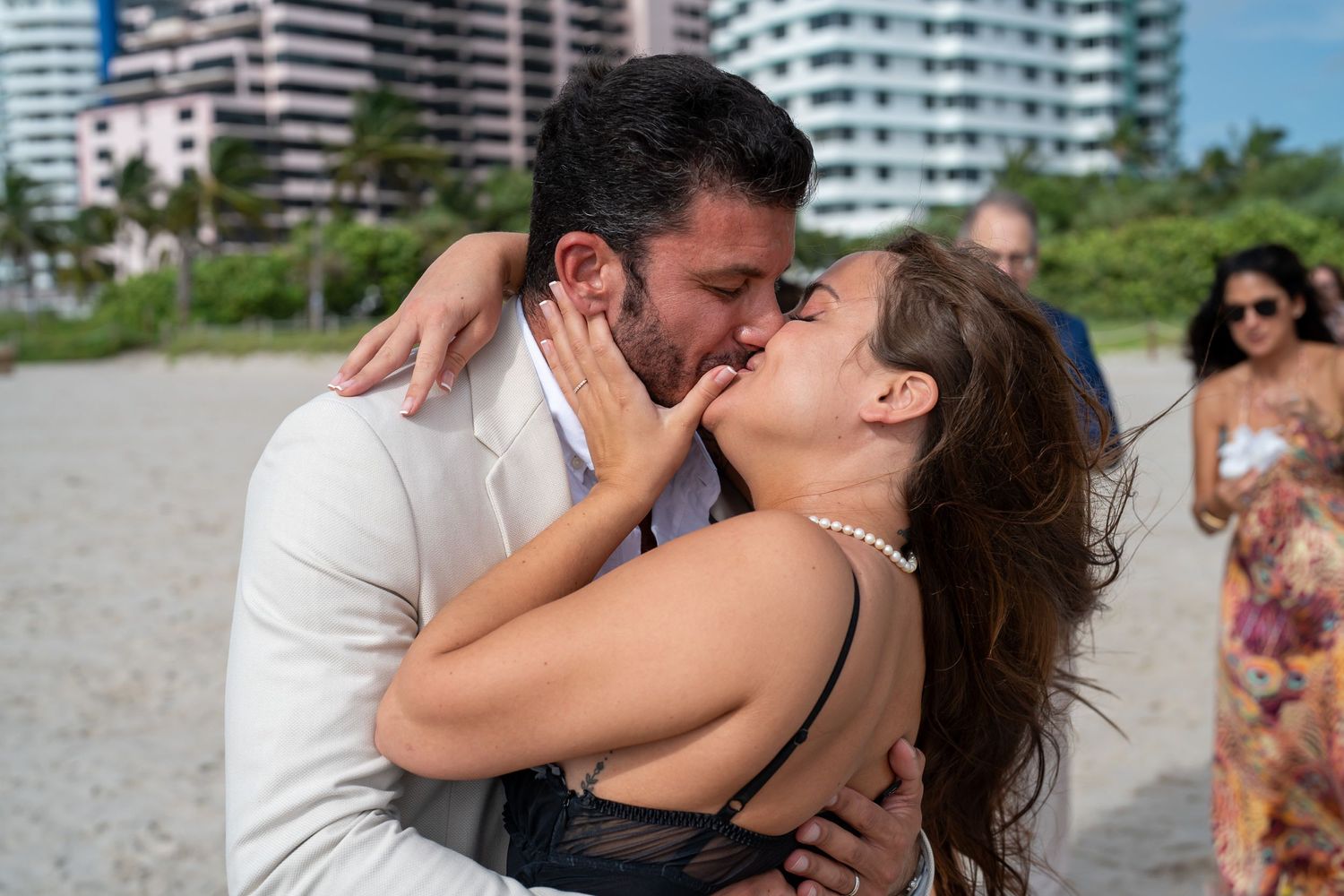 Una pareja besándose en la playa, el novio con traje blanco, con edificios altos detrás
