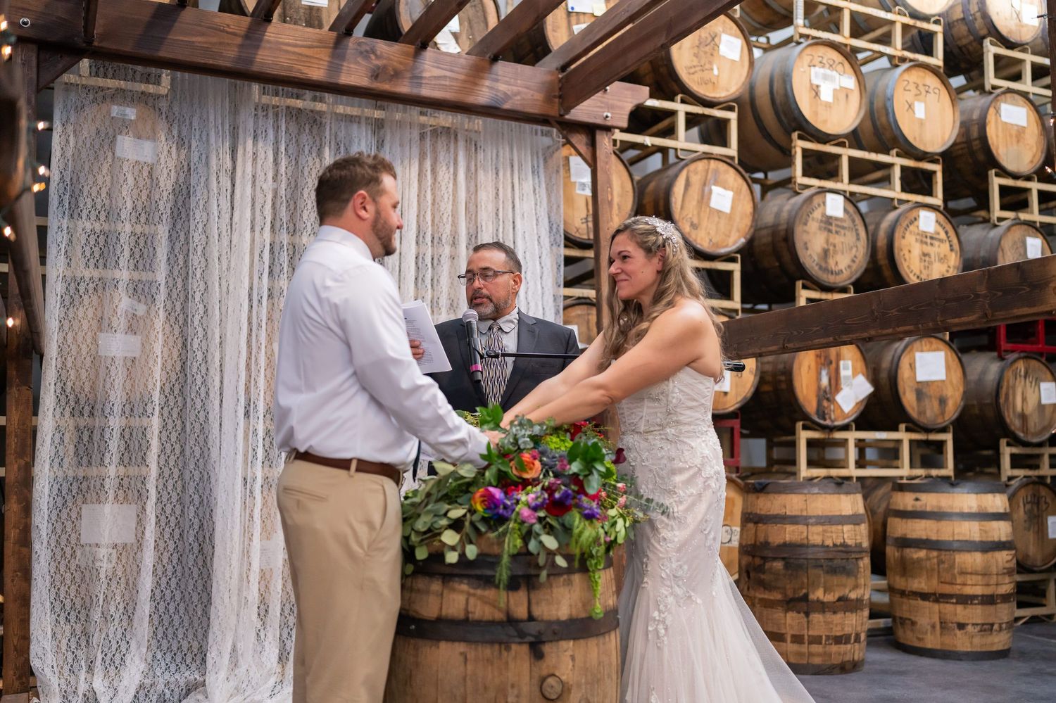 Bride and groom exchanging vows at a barrel-lined altar inside a brewery, officiant reading between them