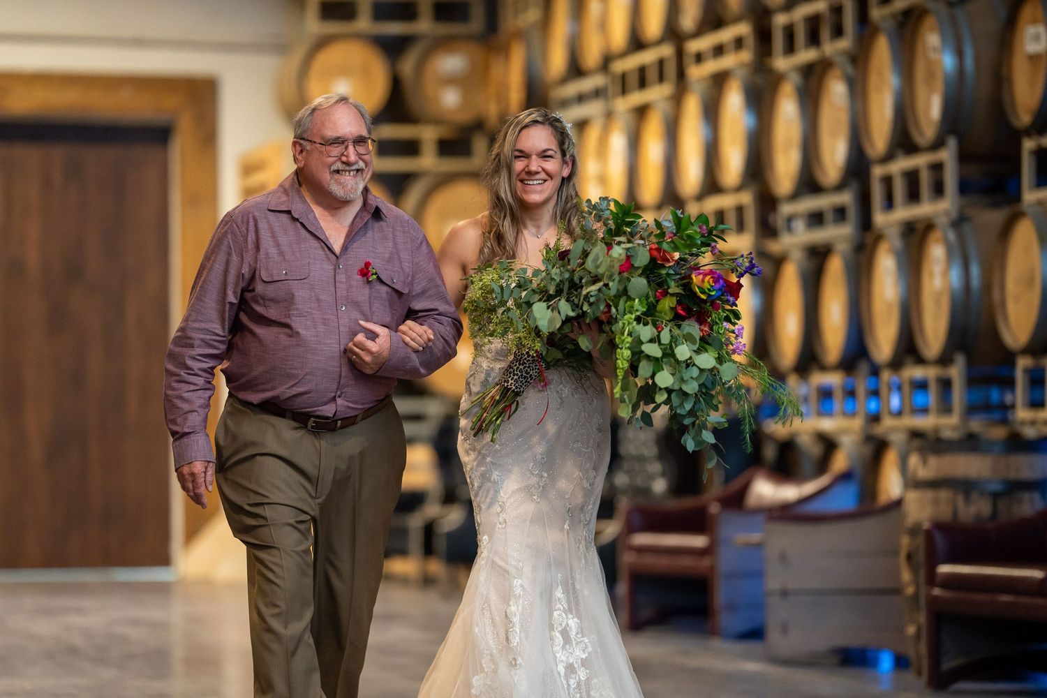 Bride's father walking her down the aisle, both laughing, in front of stacked wine barrels