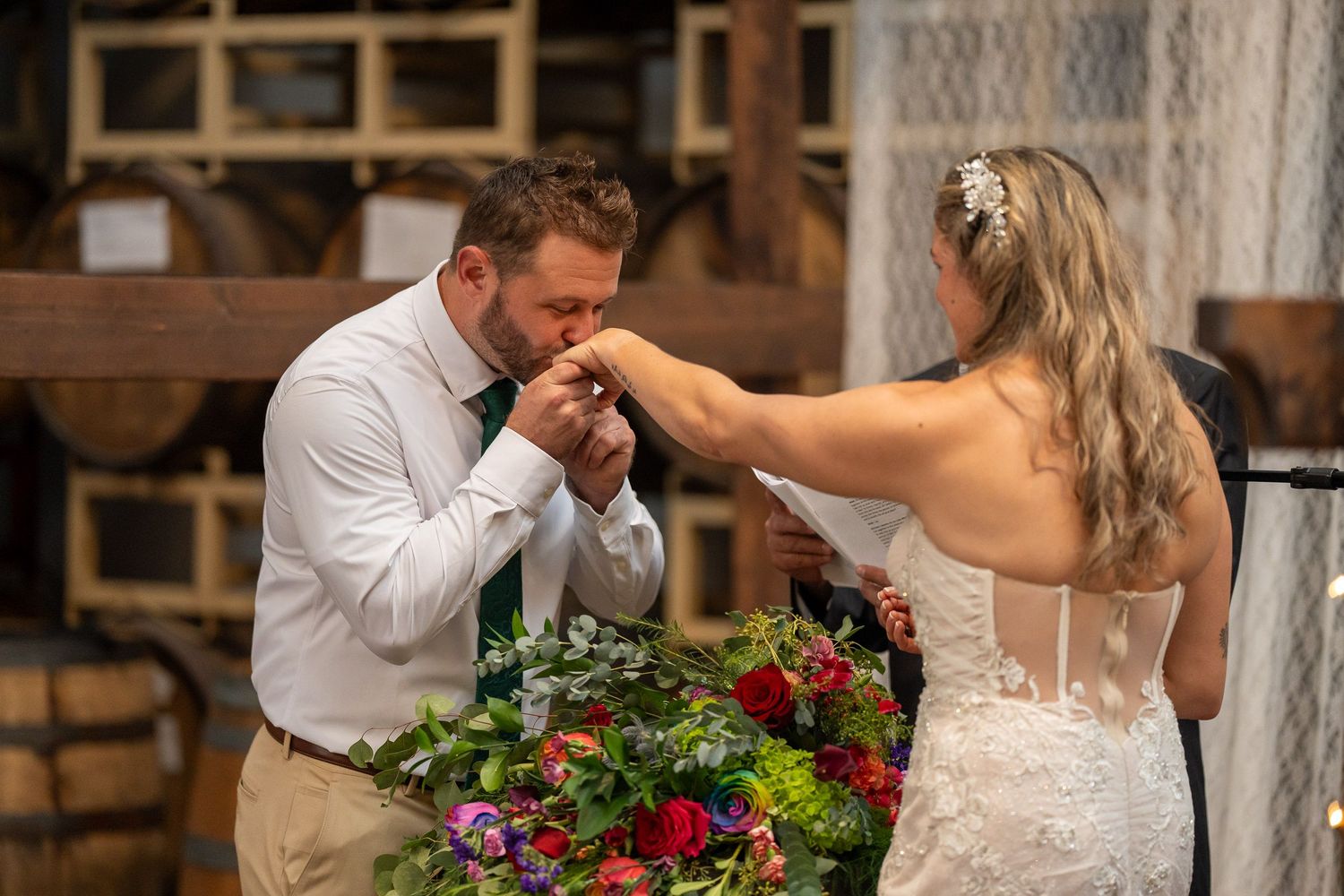 El novio besa la mano de la novia durante la ceremonia, con un ramo de flores rojas y amarillas al frente