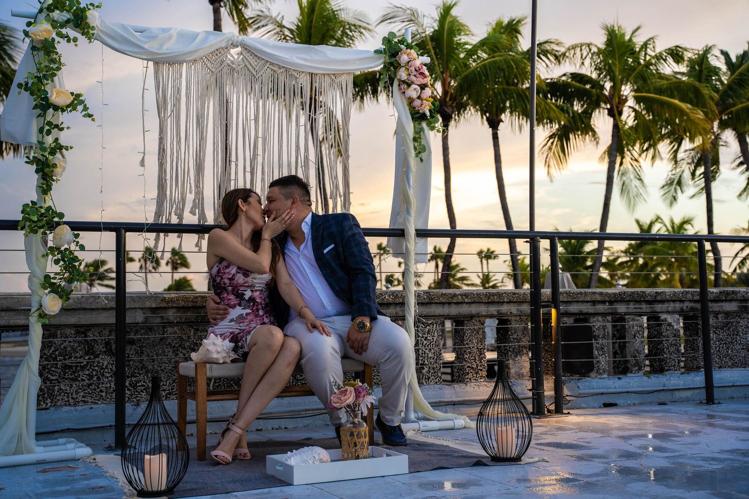 Los novios besándose bajo un arco de macramé al atardecer, con palmas a lo largo del agua detrás