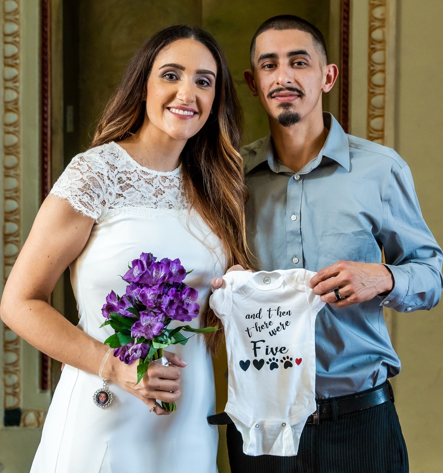 Bride and groom smiling together, holding a 'and then there were five' pregnancy announcement onesie