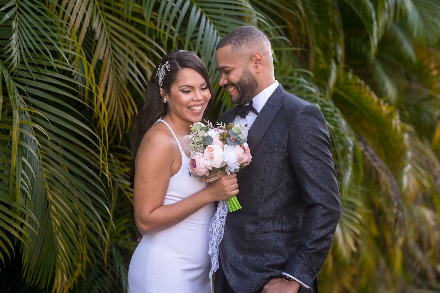 Los novios riéndose juntos frente a hojas de palma, la novia con un ramo