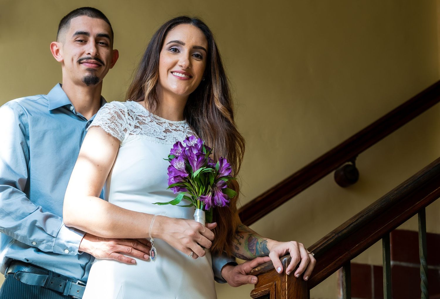 La novia con un ramo morado y el novio sonriendo junto a una escalera