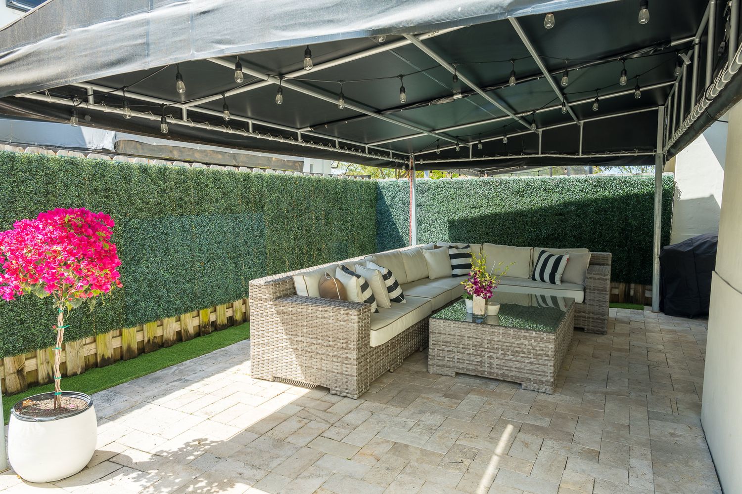 Covered outdoor patio with wicker sectional seating, string lights, and a hedge wall