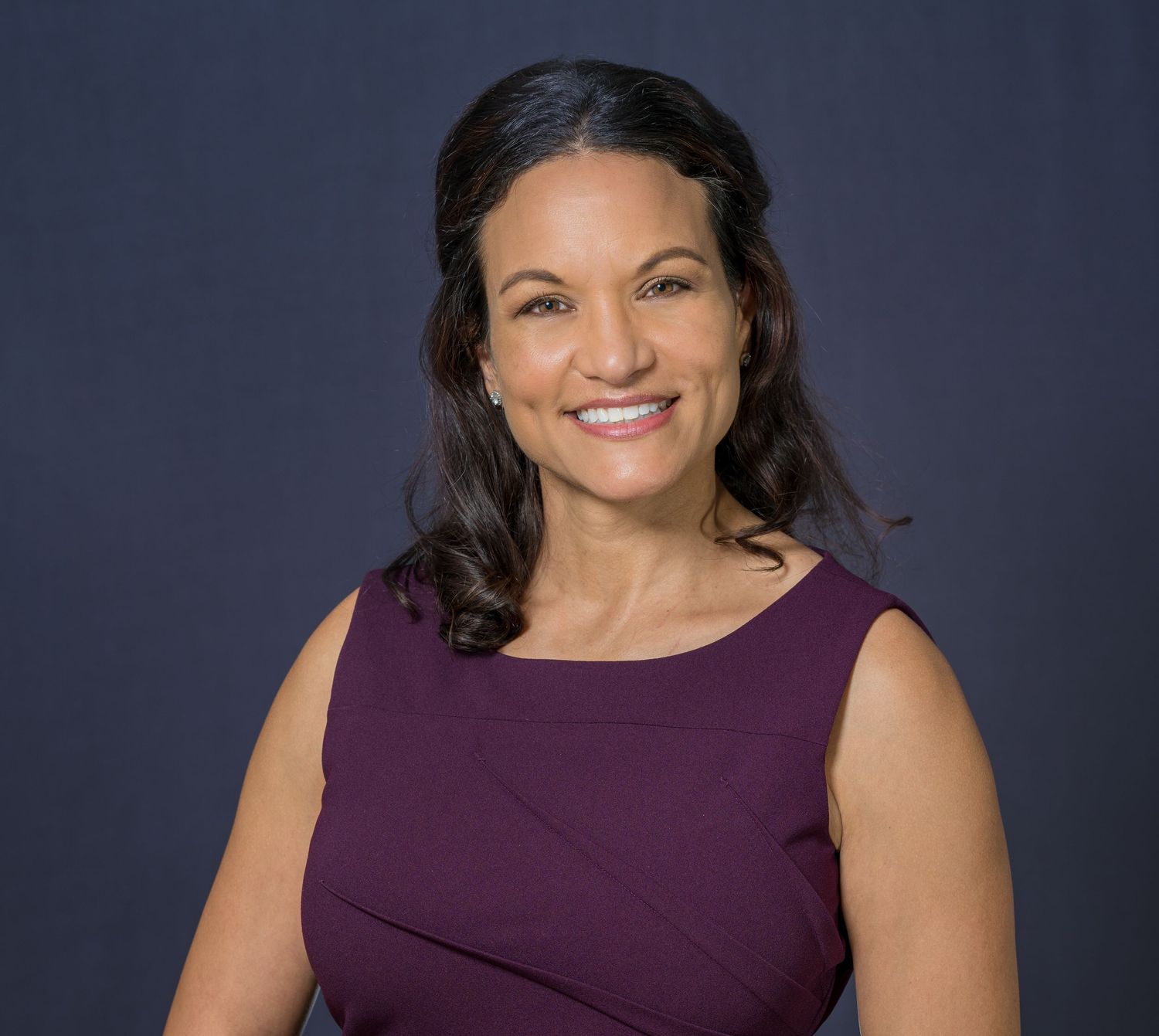 Smiling woman in a plum sleeveless top against a dark navy backdrop, studio headshot