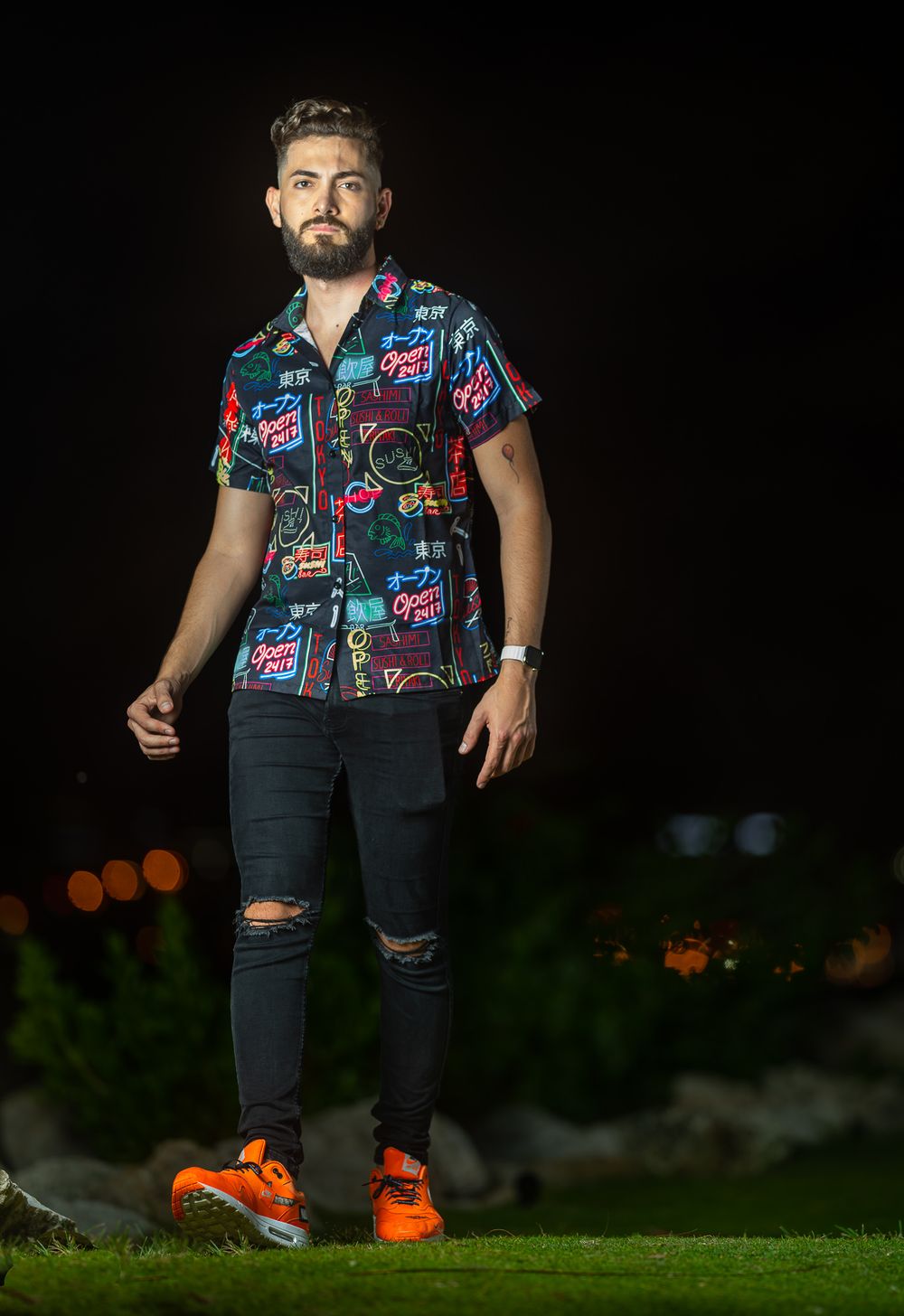 Un modelo con camisa estampada de colores y jeans negros rotos de pie frente a un fondo oscuro de noche