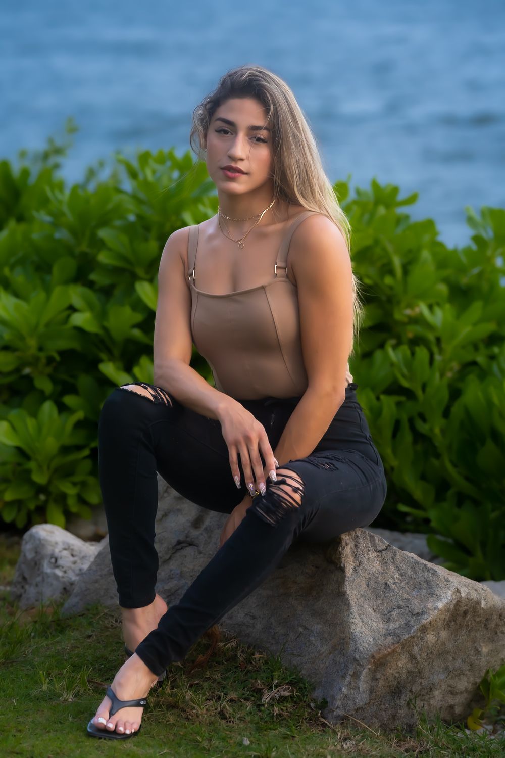 Una modelo con blusa marrón y pantalón negro sentada sobre una roca con vegetación y agua detrás al anochecer