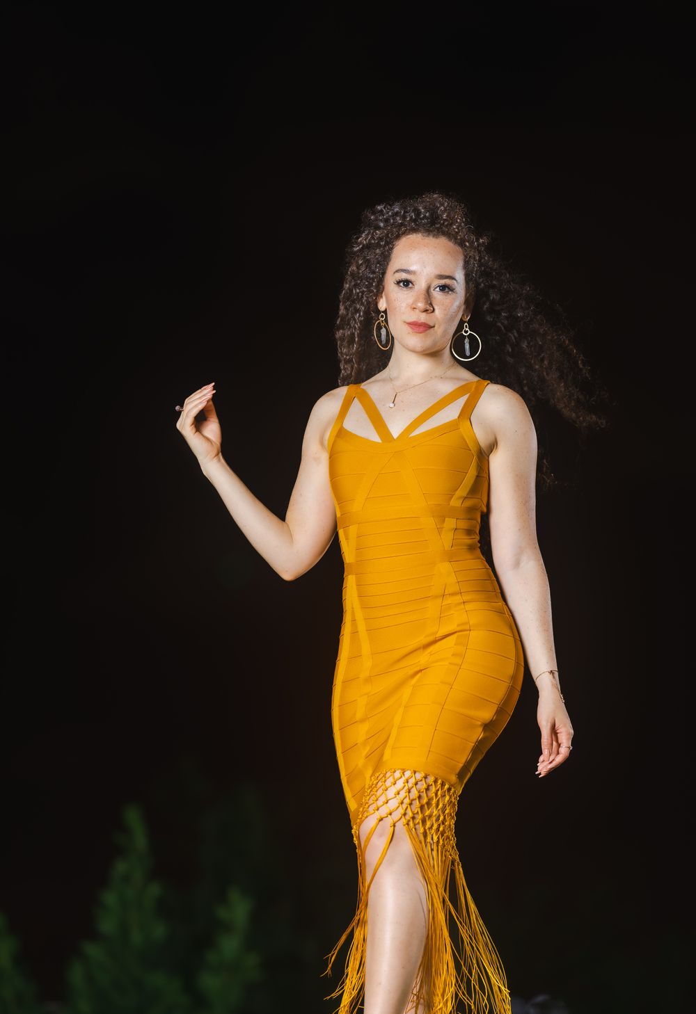 Una modelo con vestido amarillo mostaza de flecos en plena pose con un brazo extendido frente a un fondo oscuro de noche