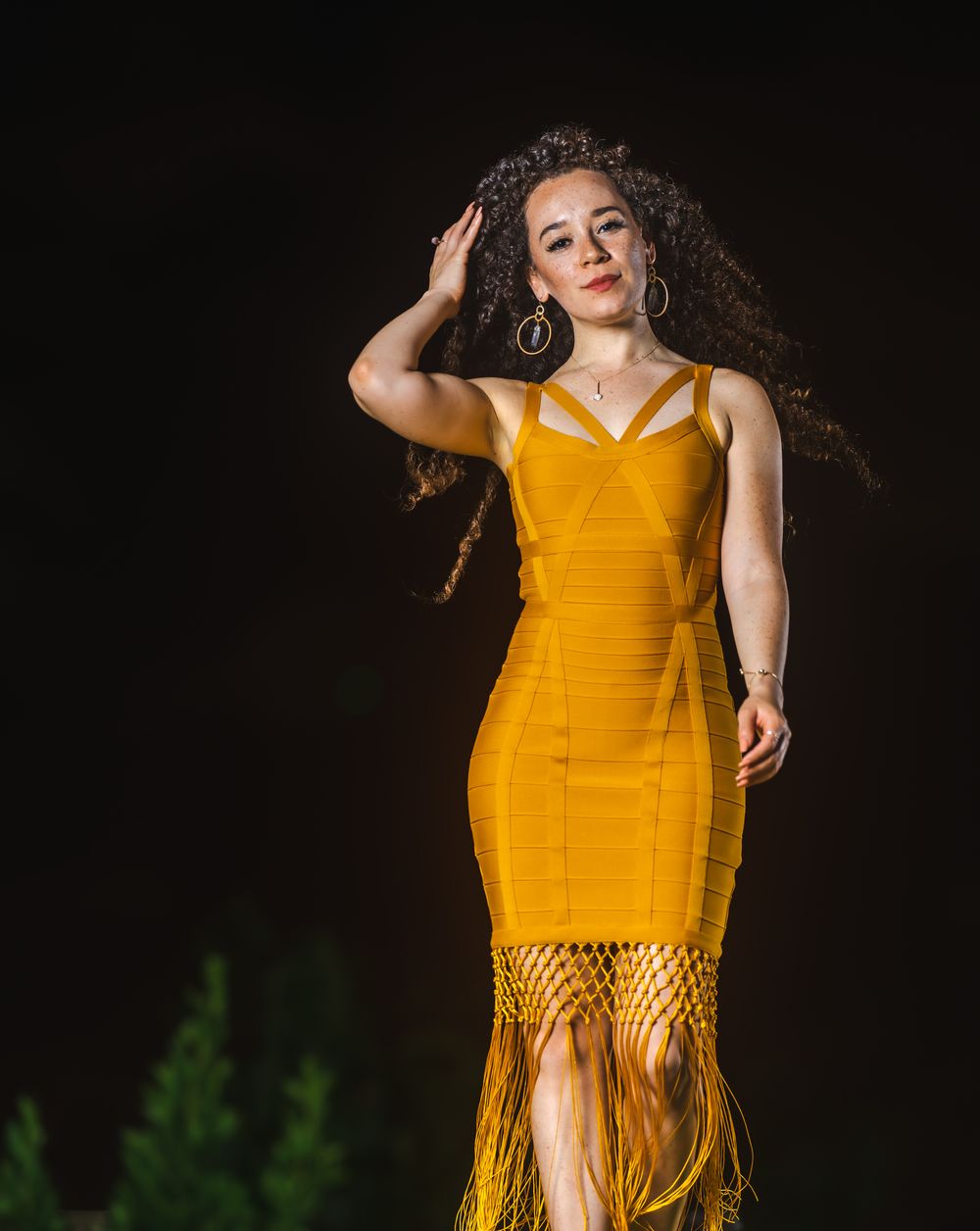 Model in a yellow fringe dress posing with her hand in her curly hair at night