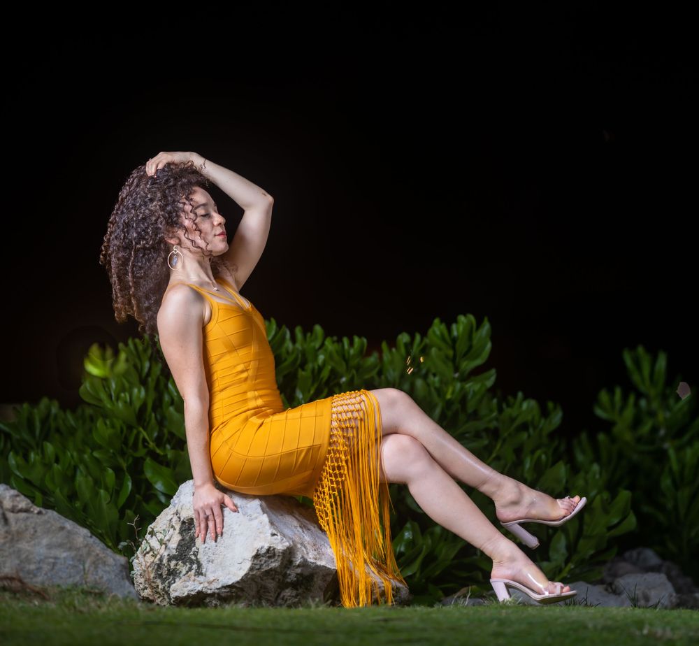 Una modelo con vestido amarillo mostaza de flecos sentada sobre una roca de noche con el pelo sobre el hombro
