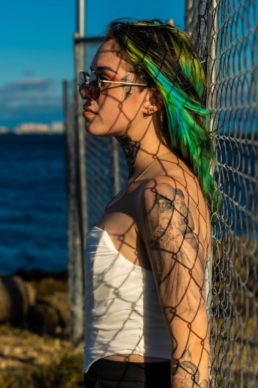 Model with colorful hair posing behind a chain-link fence near the water
