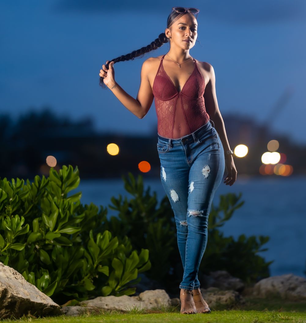 Una modelo con blusa vino y jeans rotos sostiene su trenza con las luces de la ciudad brillando detrás al anochecer