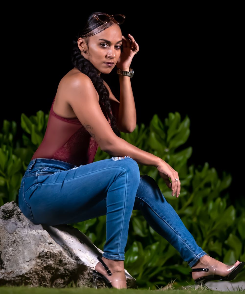 Una modelo con blusa vino y jeans sentada sobre una roca de noche con una mano en el pelo